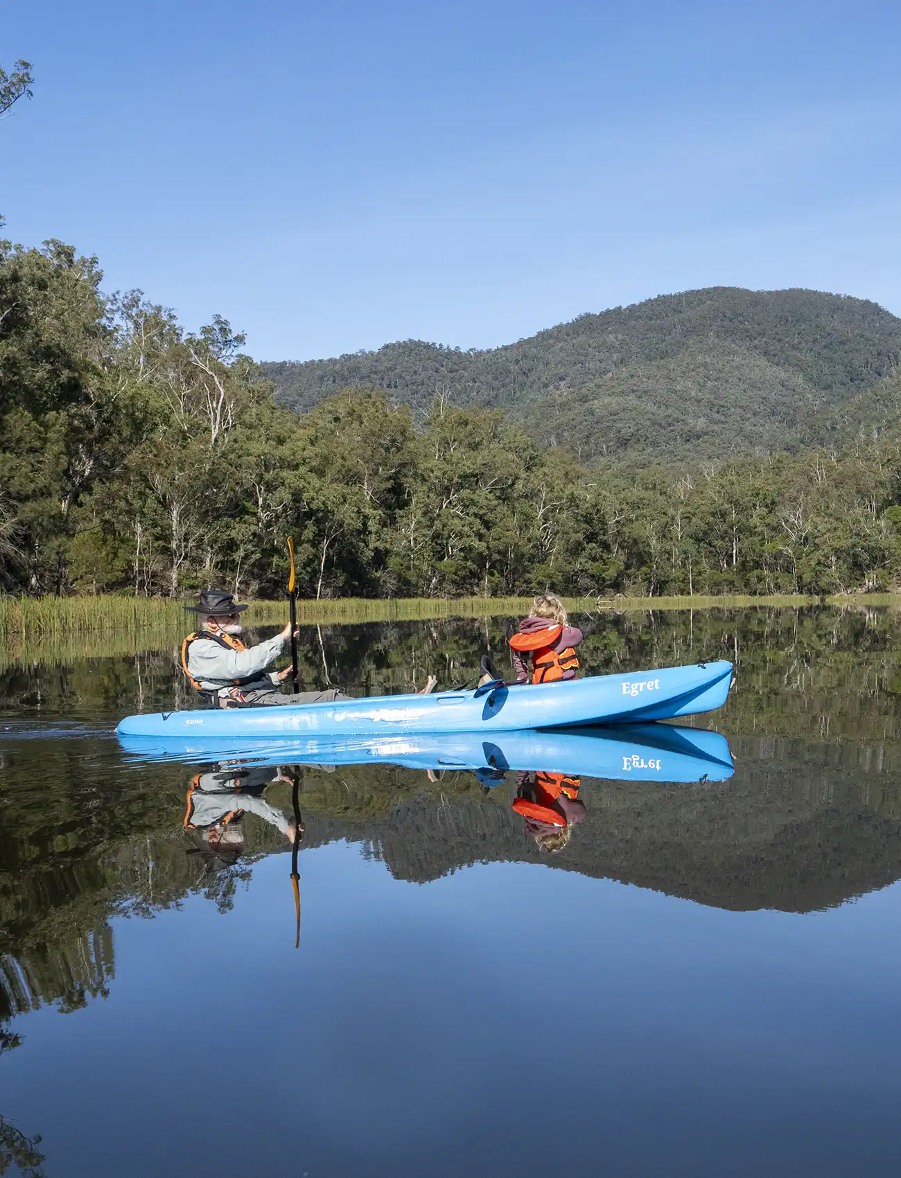 Grandfather and granddaughter kayaking on Brogo Dam, calm water, reflections, mountains in background.